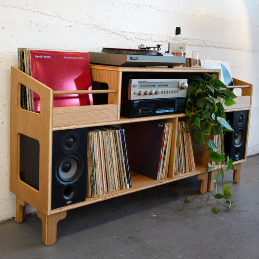 A light natural wood record shelf and media console with integrated speakers, holding vinyl records, a turntable, a cassette deck, and a potted green plant, set against a white brick wall.
