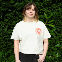 Load image into Gallery viewer, Person wearing a white t-shirt with a red logo against a green leafy background