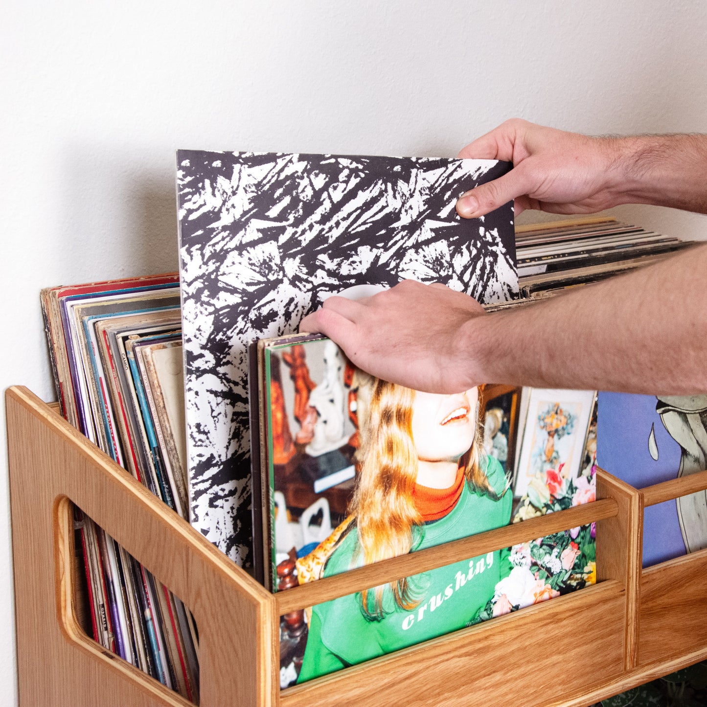 A person's hands holding a vinyl record sleeve in front of a wooden record rack filled with records, against a plain white wall.