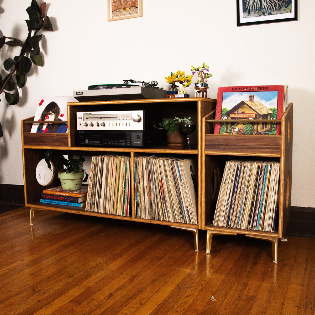 A mid-century modern wooden record shelf and media console with gold-tone legs, styled in a living room setting with vinyl records, a turntable, and framed art.