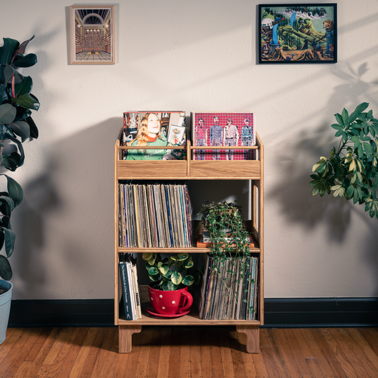 A light natural wood record shelf with three tiers stands against a beige wall on a hardwood floor, styled with vinyl records, potted plants, and framed wall art.