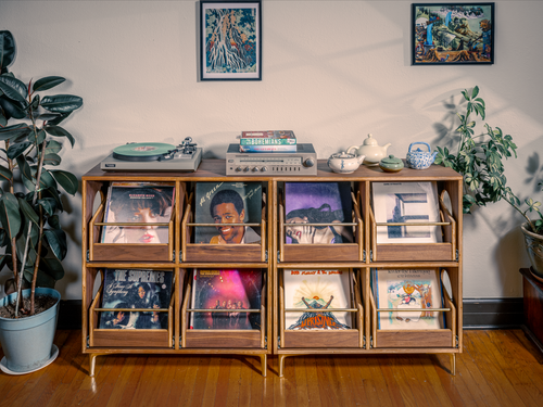 Wooden record player console with vinyl records in a room with plants and framed pictures.