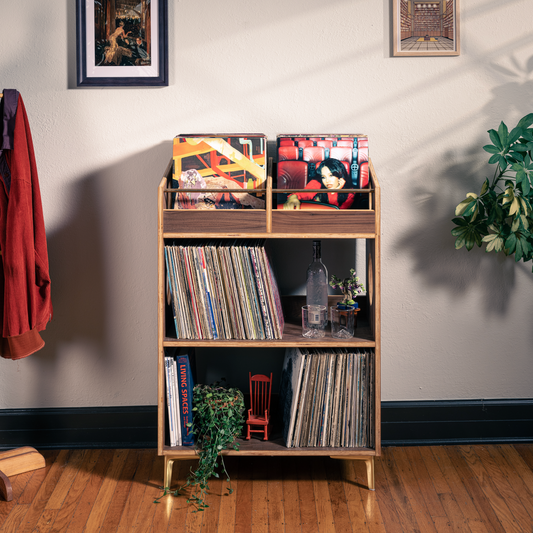 A wooden three-tier vinyl record shelf with gold legs stands in a styled room corner with a coat rack, framed art, and potted plants. The shelf is filled with records, books, and small decorative items.
