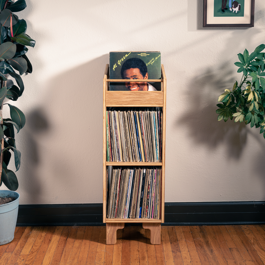 A light natural wood record rack with three tiers stands on a hardwood floor against a beige wall, holding several vinyl records and a green album cover. The room is styled with potted plants on both sides.