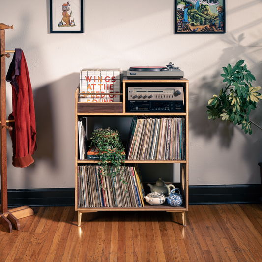 A mid-century modern wooden record shelf with gold legs stands in a styled room with hardwood floors, potted plants, a coat rack, and framed art. The shelf is styled with vinyl records, a turntable, and decorative items.