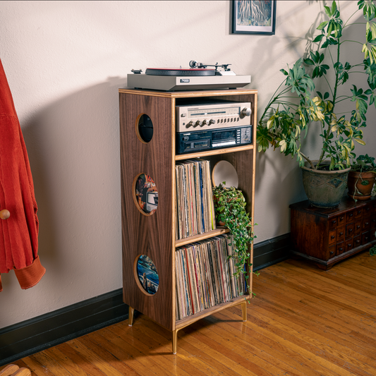 A tall walnut wood record cabinet with circular cutouts and gold legs, styled in a room with a record player, vinyl records, and houseplants.