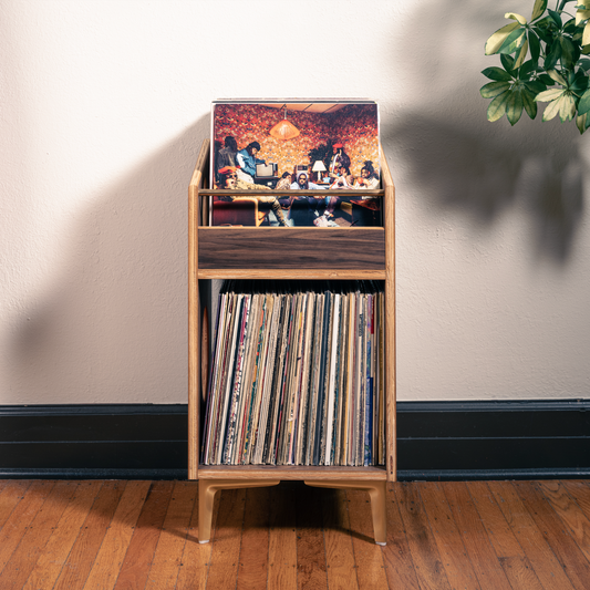 A mid-century modern wooden record rack stand holding vinyl records and a record sleeve, set against a cream wall with a plant and red garment visible in a styled room setting.