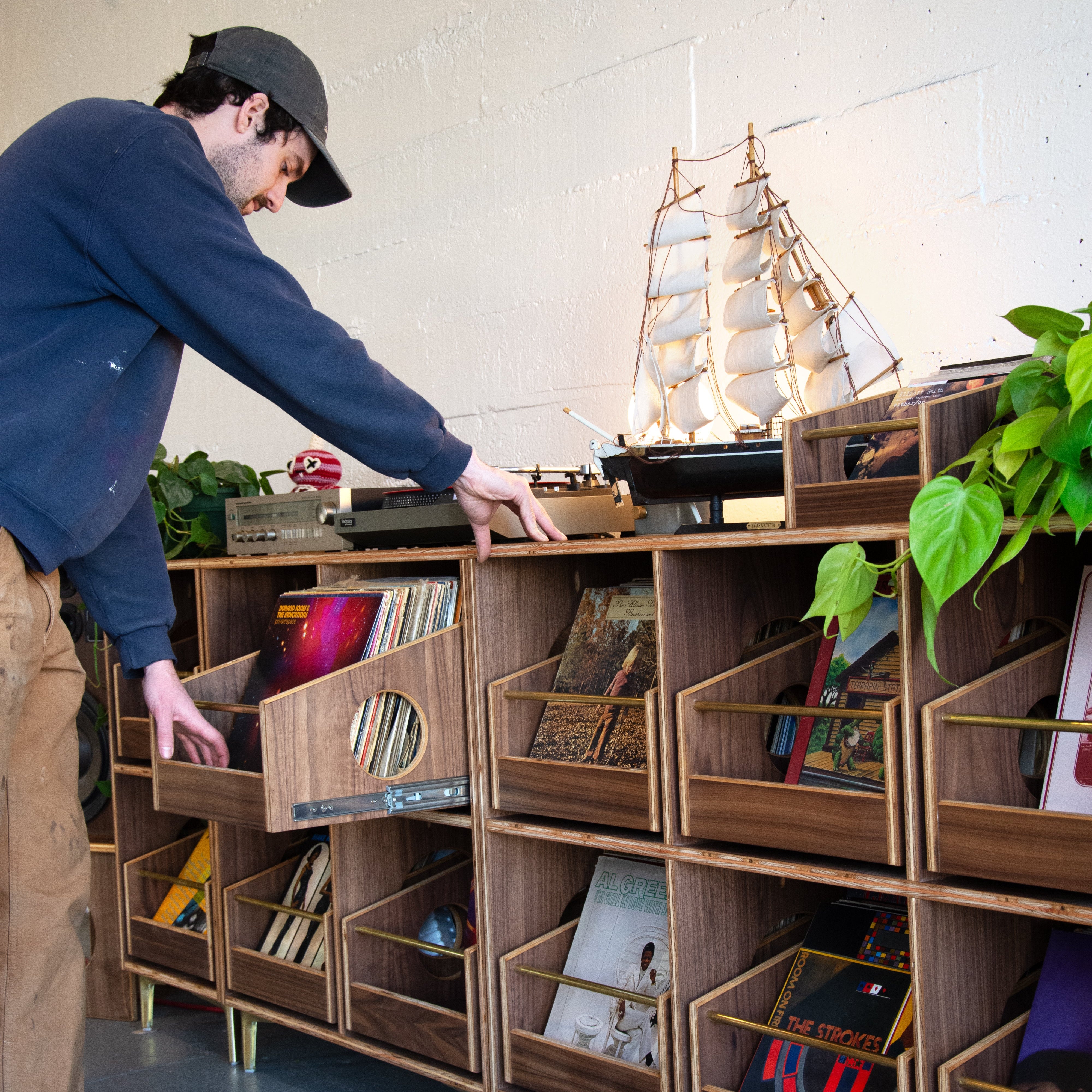 Person organizing records on a wooden shelf with a model ship and plants.