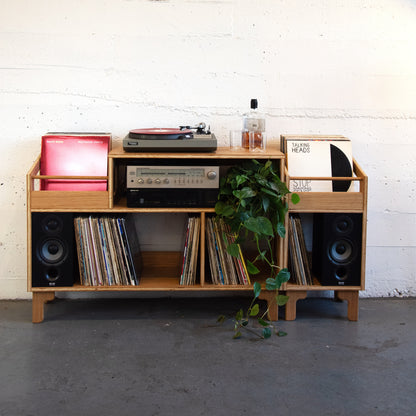 A mid-century modern wooden record rack console with integrated speakers, vinyl records, a turntable, and a plant, set against a white brick wall and concrete floor.