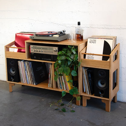 A light natural wood vinyl record rack with integrated shelves, side bins, and speaker cutouts, styled with a turntable, speakers, records, and a trailing green plant against a white brick wall.