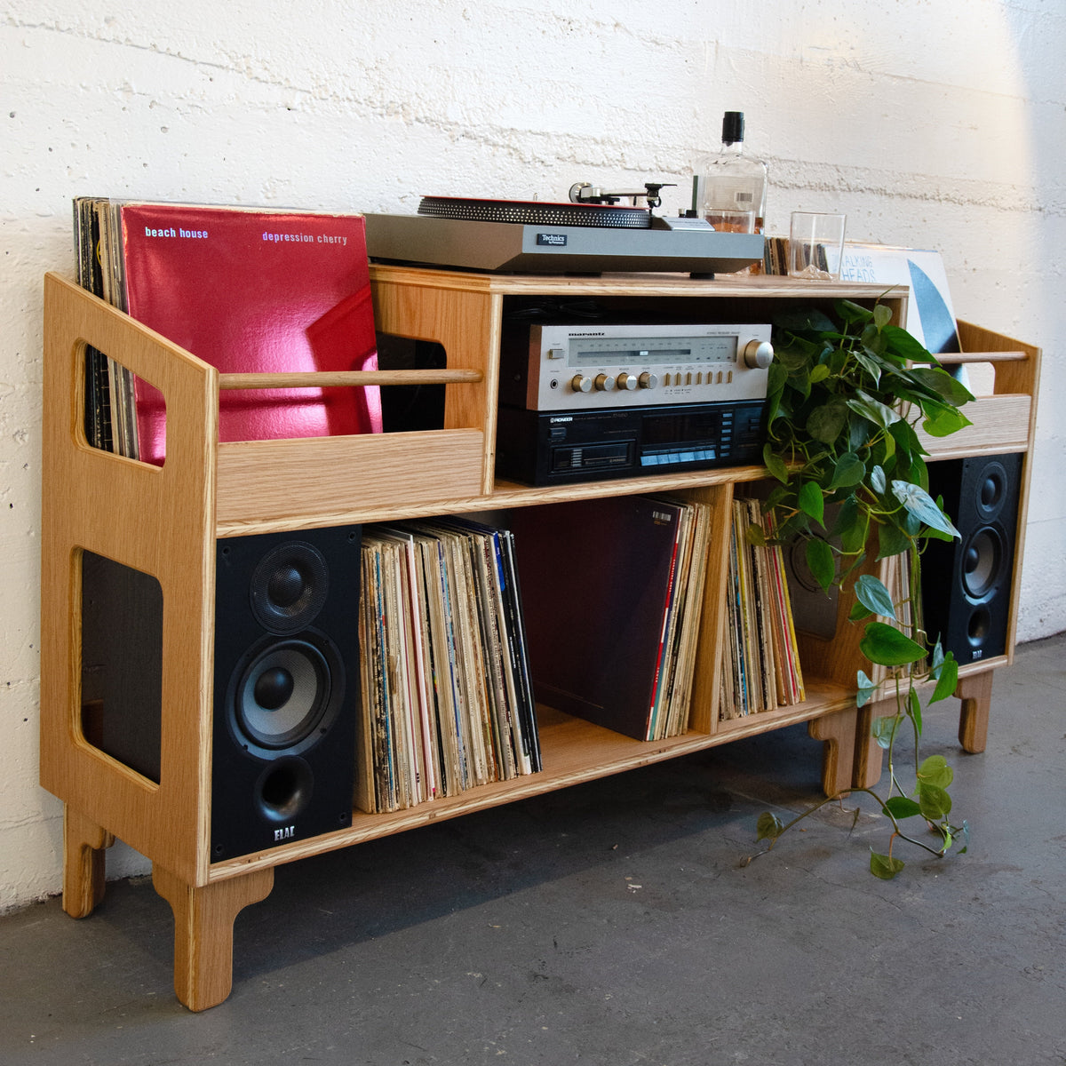 A light natural wood record shelf and media console with integrated speakers, holding vinyl records, a turntable, a cassette deck, and a potted green plant, set against a white brick wall.