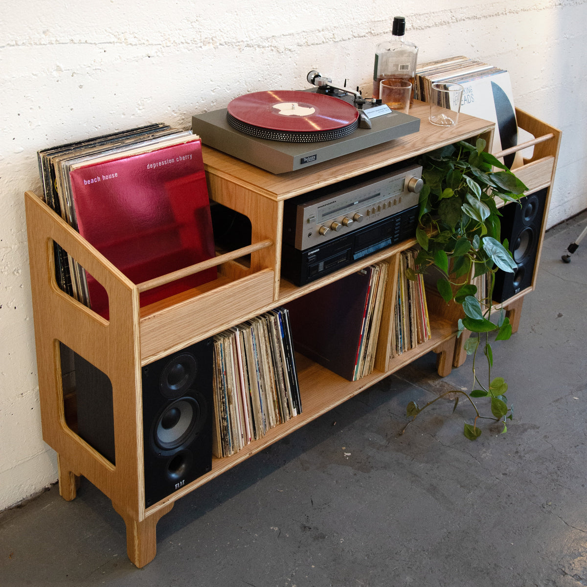 A light natural wood record player media console styled with a turntable, vinyl records, speakers, and a potted green plant, set against a white painted brick wall.