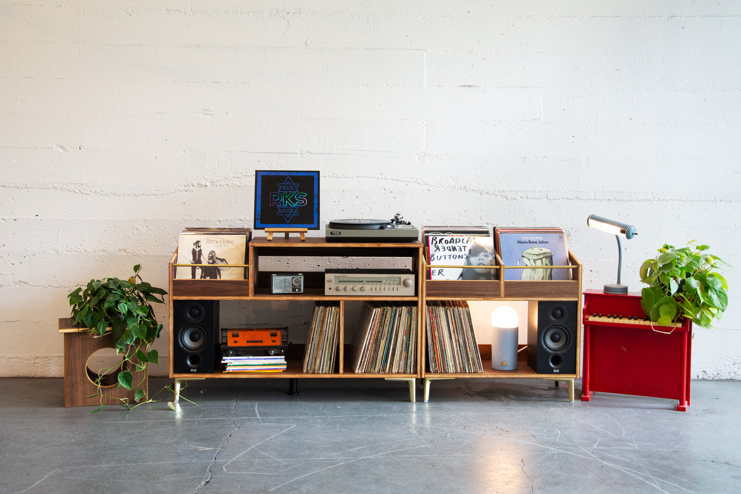 Wooden record player console with vinyl records and speakers in a room with a white wall and gray floor.