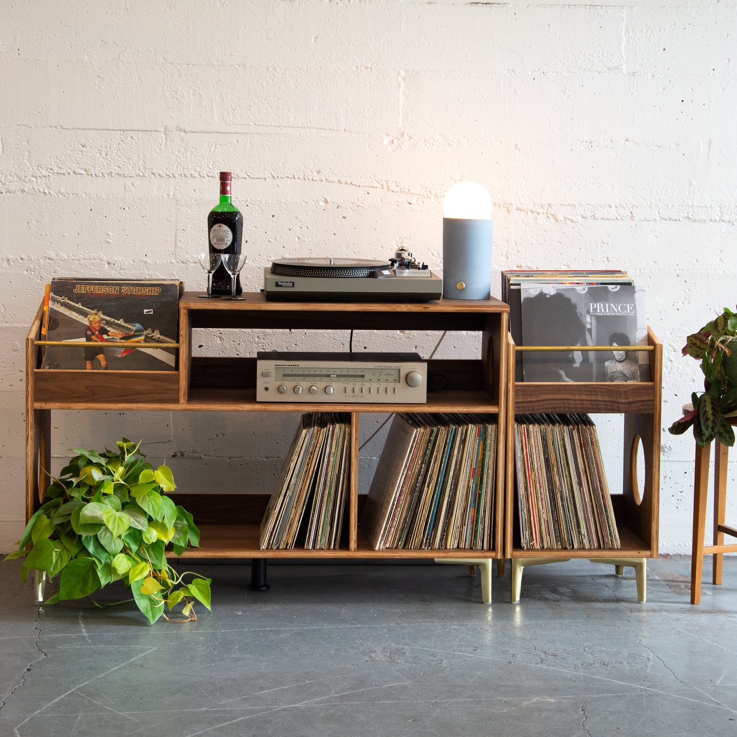 Wooden record player console with vinyl records and a plant in a minimalistic room.