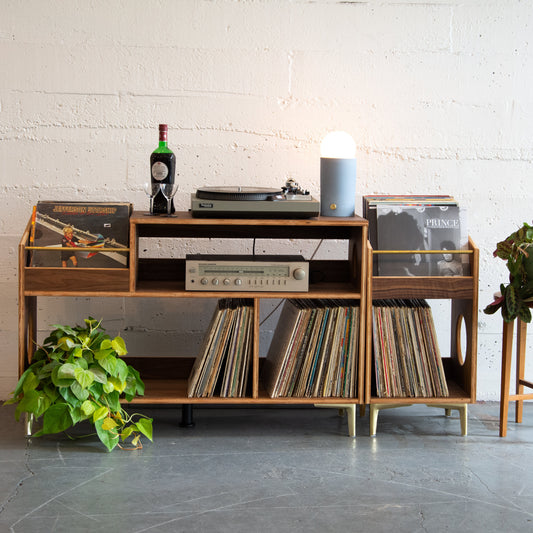 Wooden record player console with vinyl records and a plant in a minimalistic room.