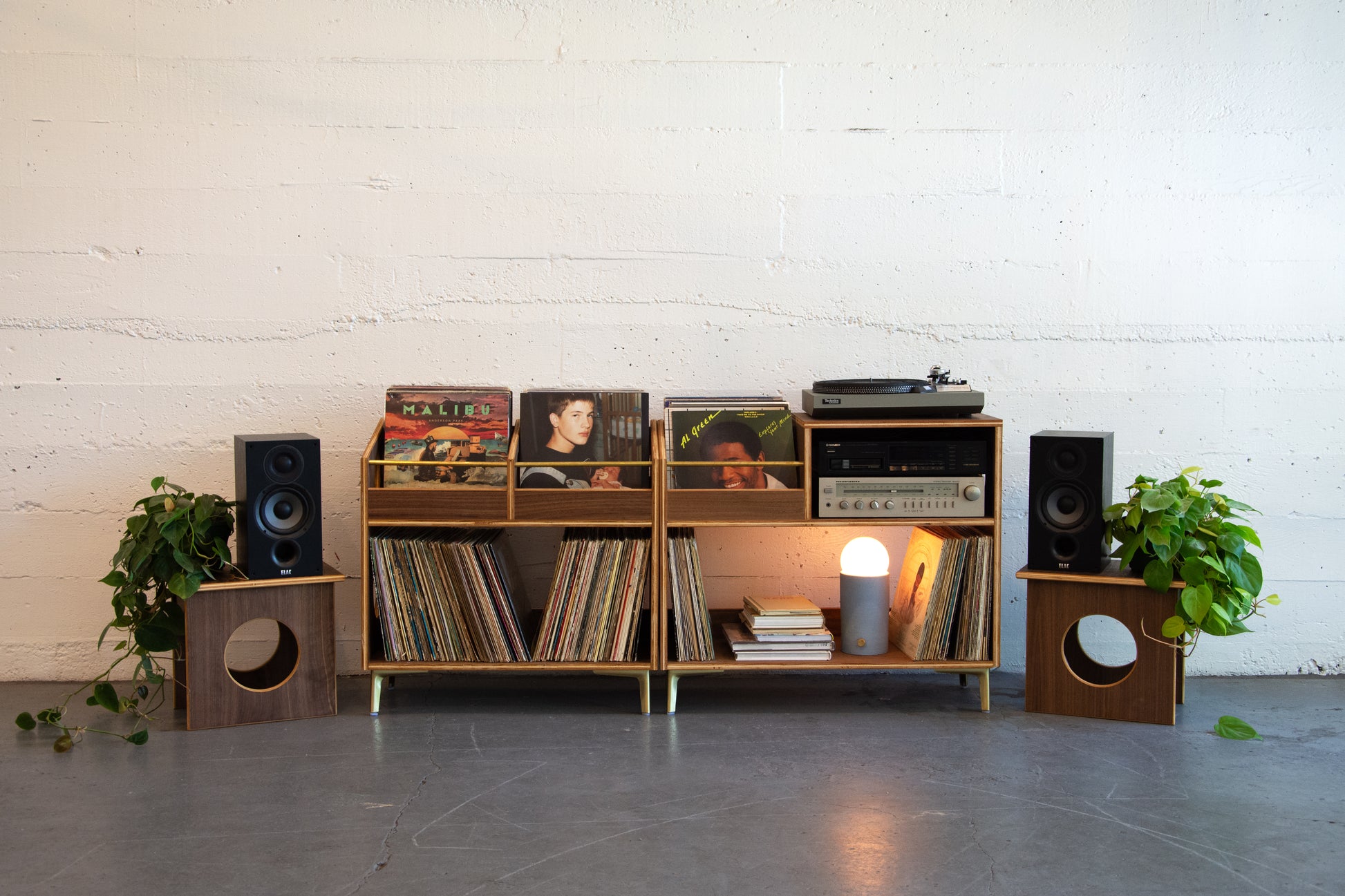 Wooden record player console with vinyl records, speakers, and a lamp against a white brick wall.