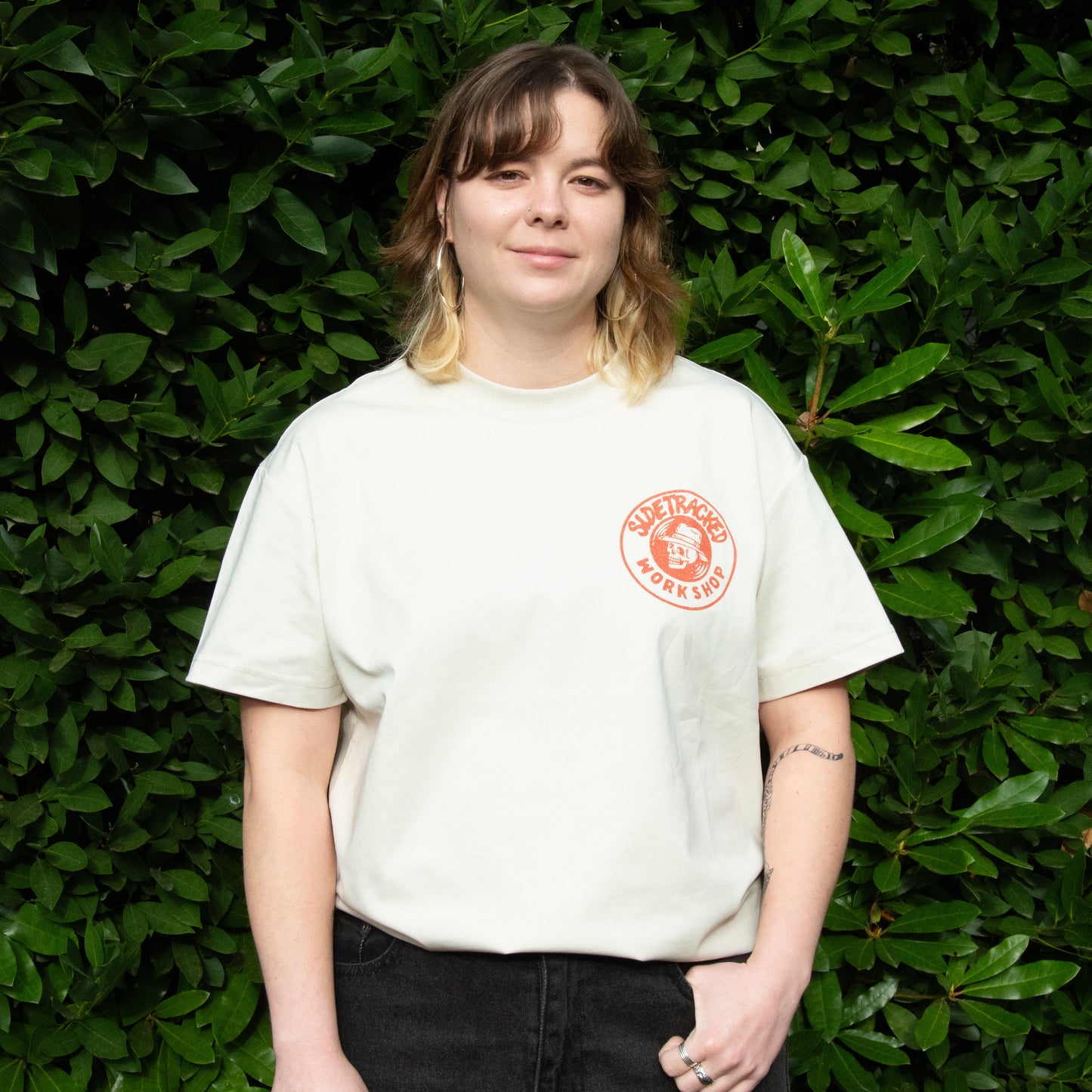 Person wearing a white t-shirt with a red logo against a green leafy background