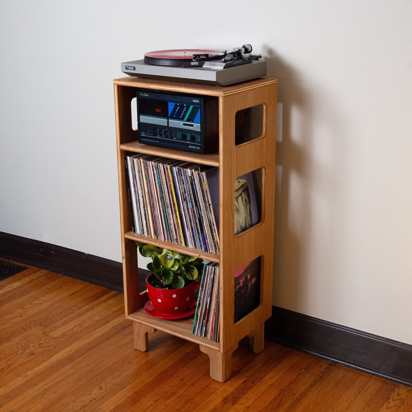 A light natural wood record player stand in a home setting, holding a turntable, a stereo receiver, vinyl records, and a small potted plant, set against a plain off-white wall and wooden floor.