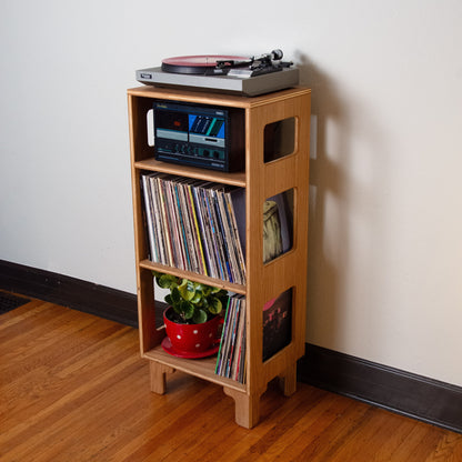 A light natural wood record player stand in a home setting, holding a turntable, a stereo receiver, vinyl records, and a small potted plant, set against a plain off-white wall and wooden floor.