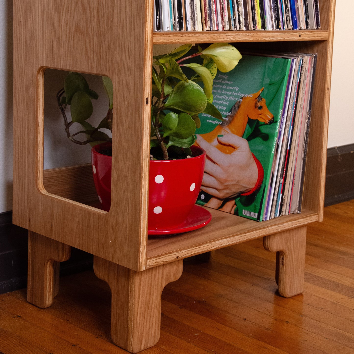 A light natural wood record shelf with cutout side panels, holding vinyl records and a potted green plant in a red polka-dot pot on a wooden floor.