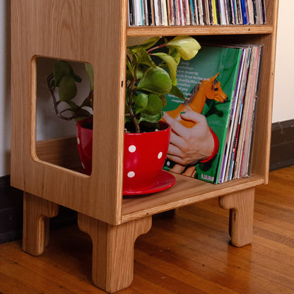 A light natural wood record shelf with cutout side panels, holding vinyl records and a potted green plant in a red polka-dot pot on a wooden floor.