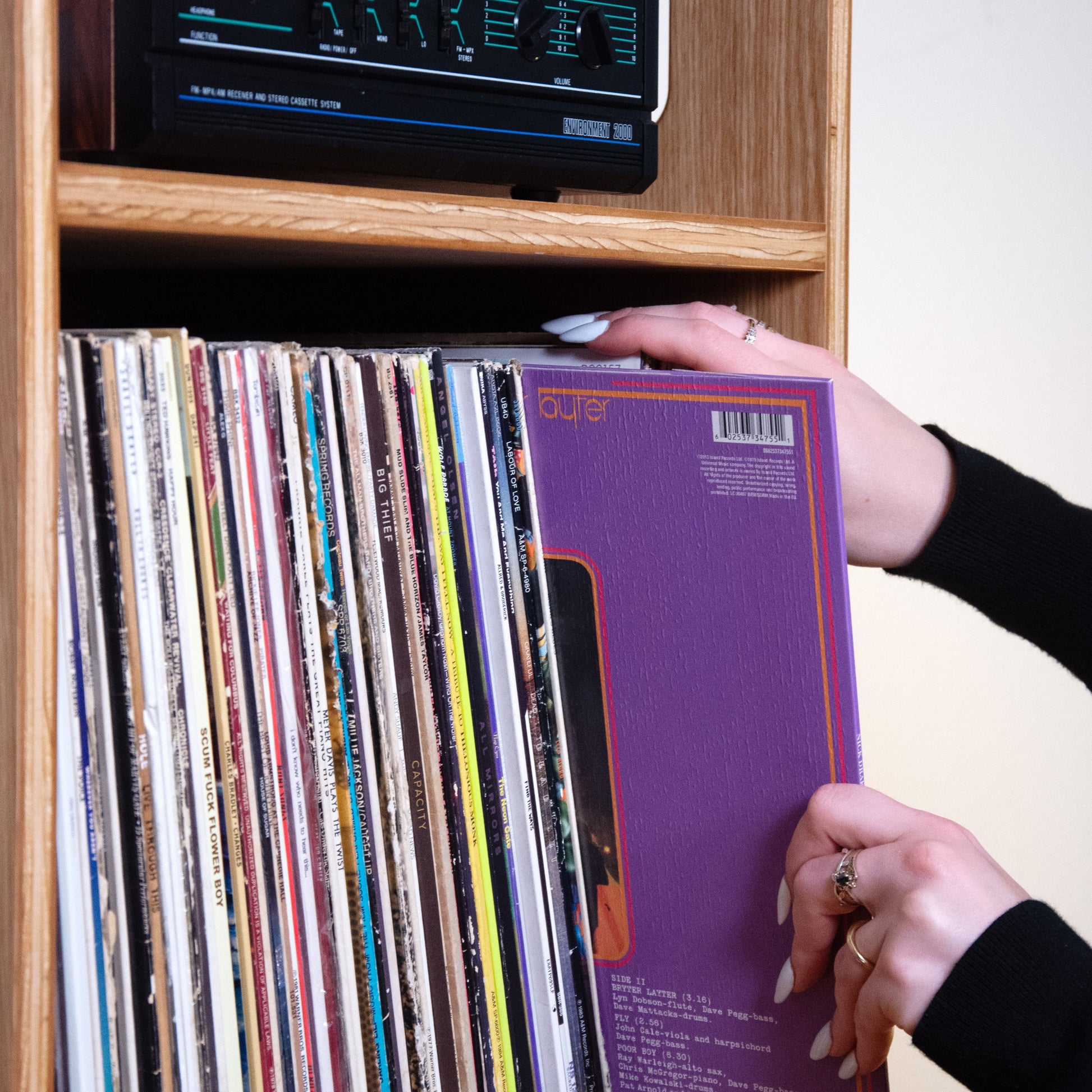 A person's hands are shown pulling a purple vinyl record from a wooden shelf filled with records, with a cassette player visible above.