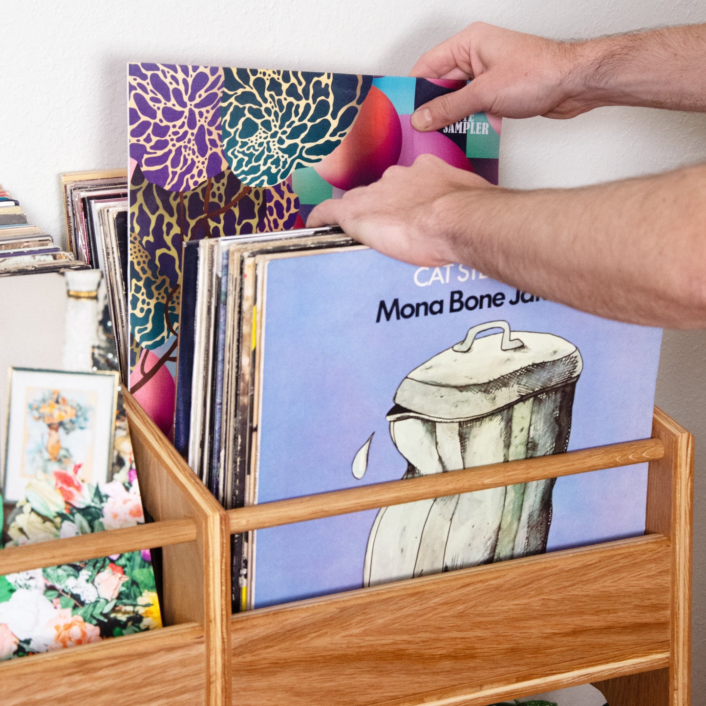 A person's hands holding a vinyl record in a wooden record rack filled with records against a white wall.
