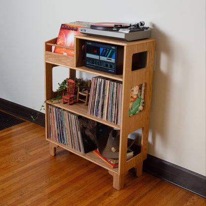 A light natural wood record shelf with three tiers, styled with vinyl records, a turntable on top, and small decorative items, set against a plain white wall on a hardwood floor.