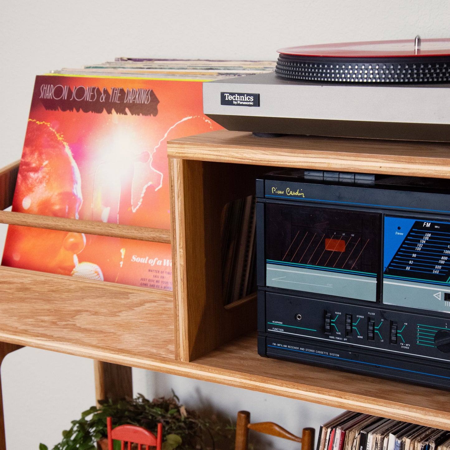 A wooden record shelf holding vinyl records, a turntable, a cassette deck, and small decorative chairs, photographed in a real interior setting.