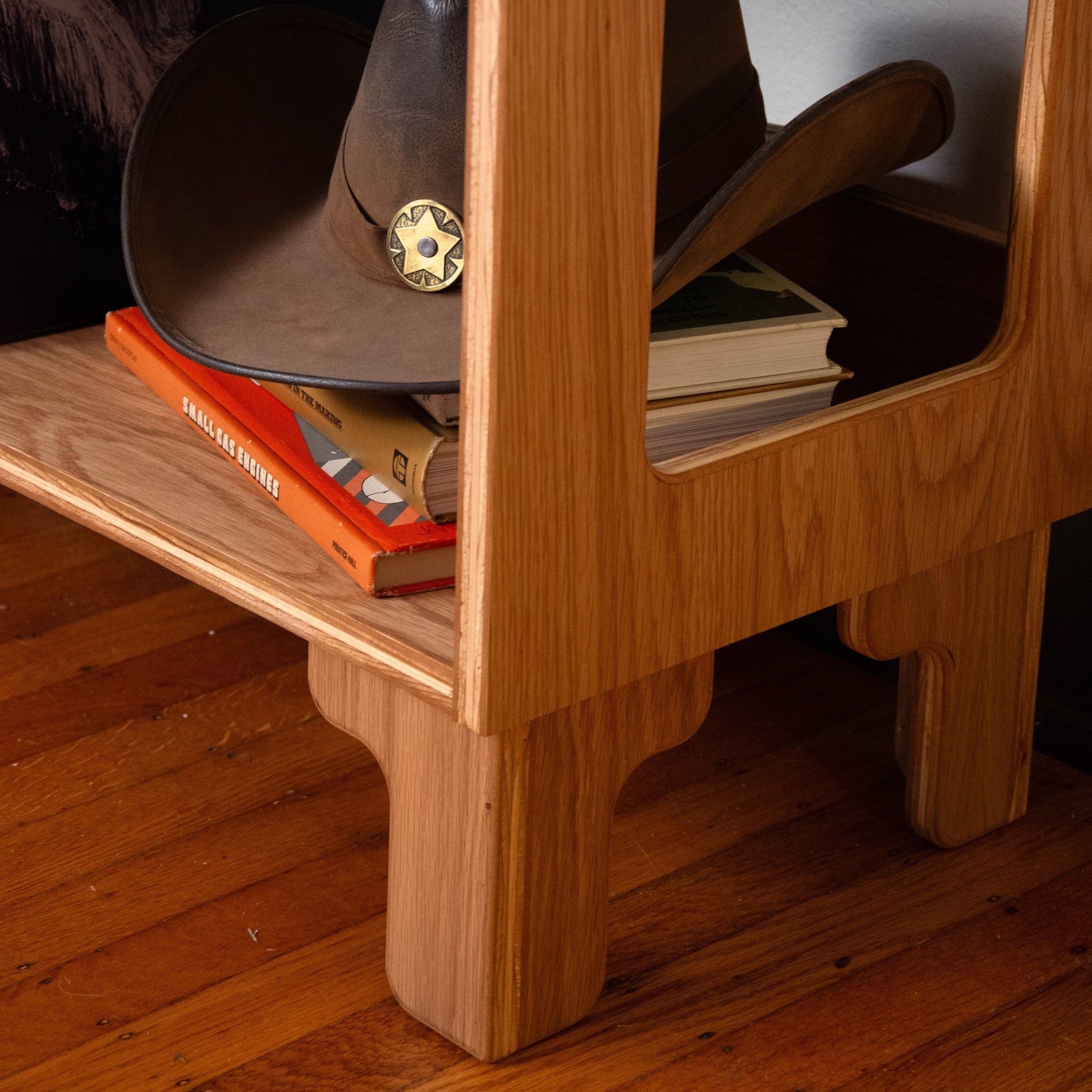A close-up of a light brown wooden side table or shelf with a cutout window, holding a dark brown cowboy hat with a gold sheriff star badge and orange books, set on a hardwood floor.