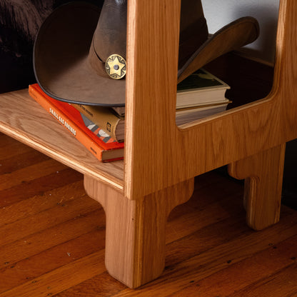 A close-up of a light brown wooden side table or shelf with a cutout window, holding a dark brown cowboy hat with a gold sheriff star badge and orange books, set on a hardwood floor.