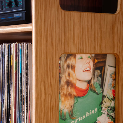 A close-up of a wooden record player or speaker cabinet with a square inset photo of a woman in a green sweatshirt that reads 'crushing'. Vinyl records are visible on a shelf to the left.