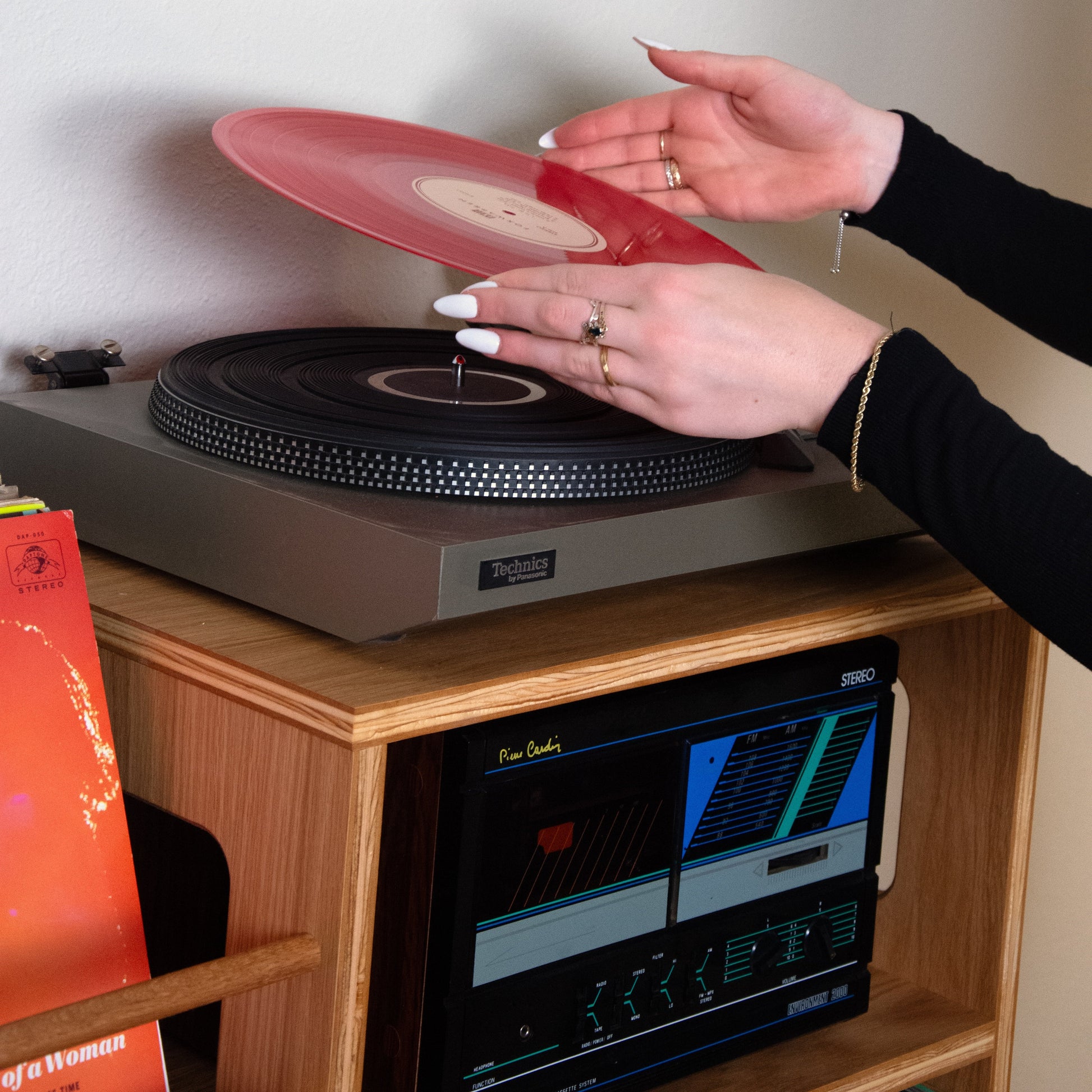A person's hands lift a translucent red vinyl record onto a black turntable sitting on a wooden cabinet, with a vintage radio below.