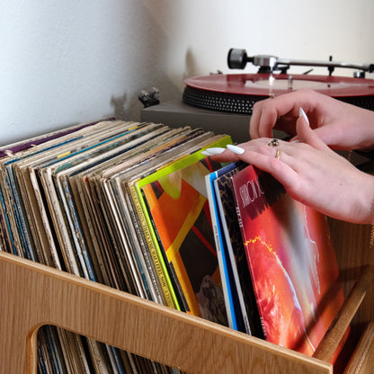 A wooden record bin filled with vinyl records, with a person's hands reaching for a record in the front. A turntable is visible in the background against a plain wall.