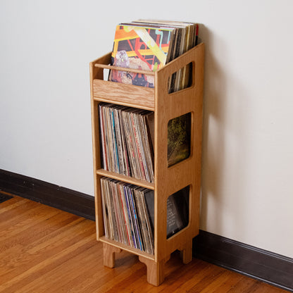 A light natural wood record rack with open shelves and side windows, filled with vinyl records, standing on a hardwood floor against a plain off-white wall.