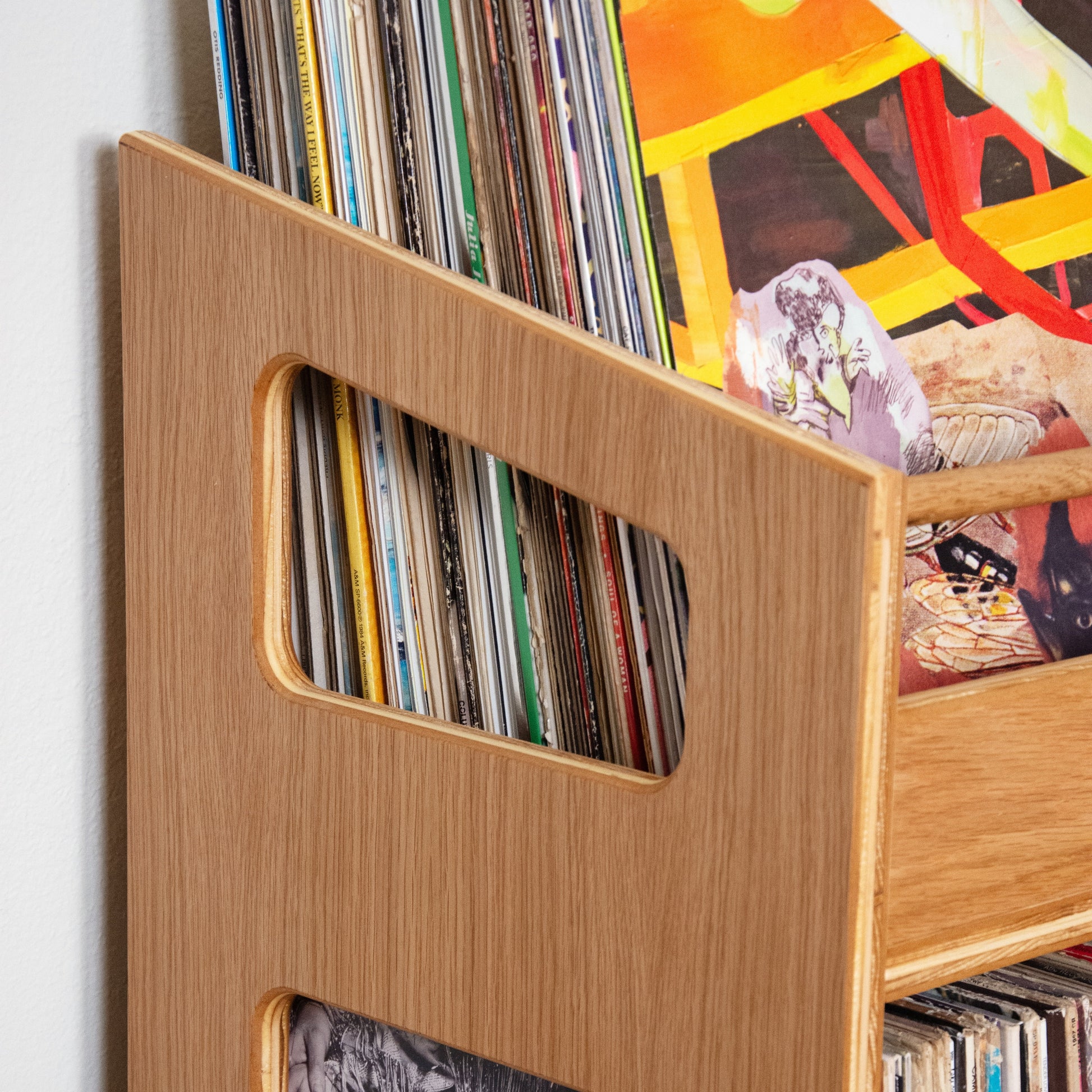 Close-up of a light natural wood record rack holding vinyl records and a framed black-and-white print, with colorful artwork visible behind it.