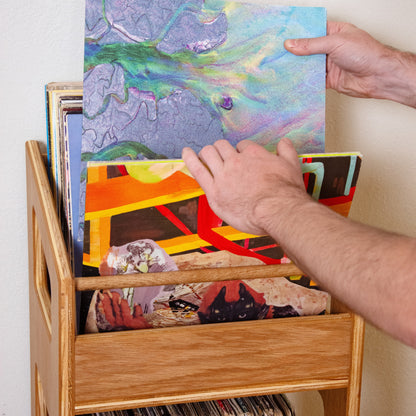 A person's hands holding up a colorful abstract art print or card above a wooden record rack filled with vinyl records.