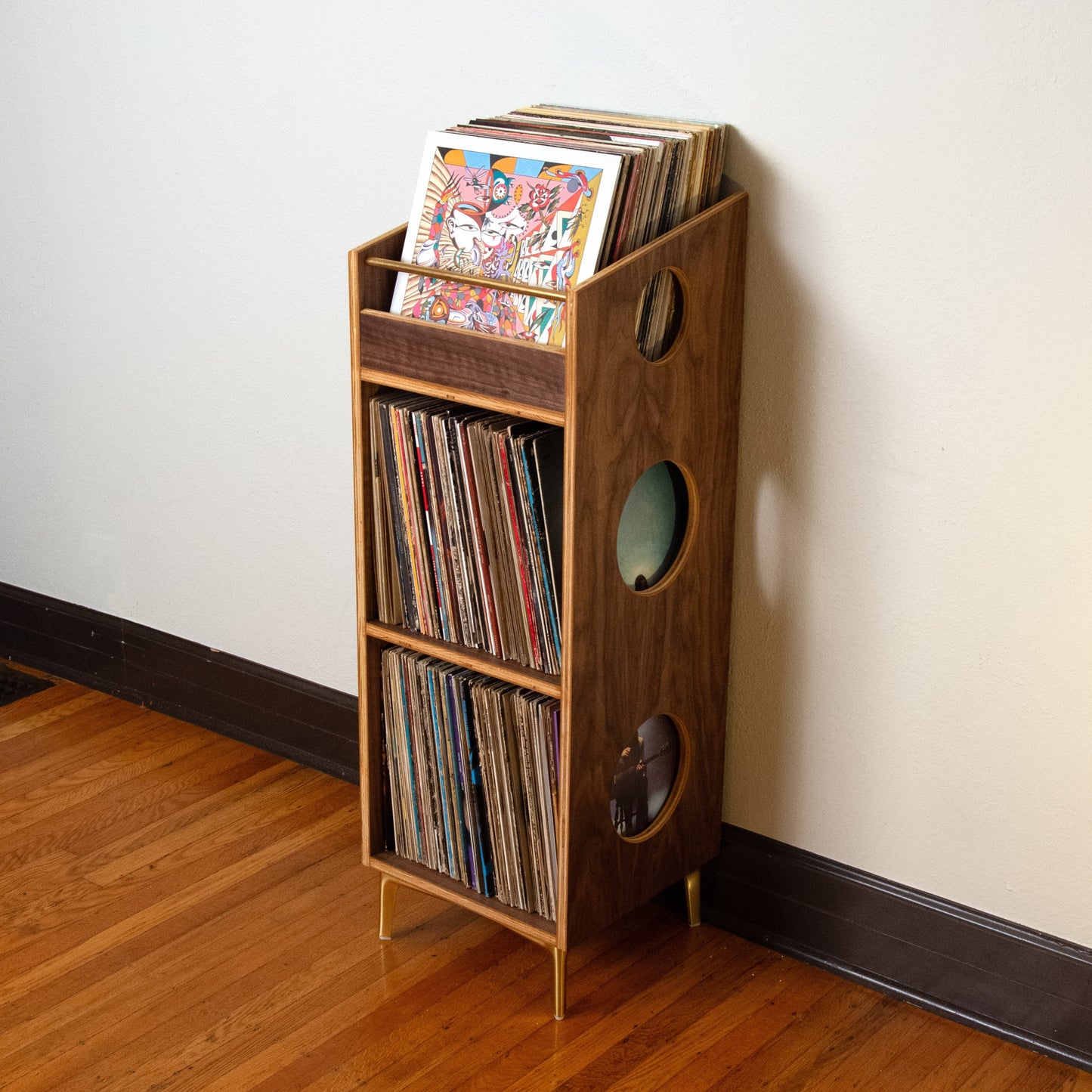 A wooden record rack shelf with circular cutouts, filled with vinyl records, standing on a hardwood floor against a plain off-white wall.