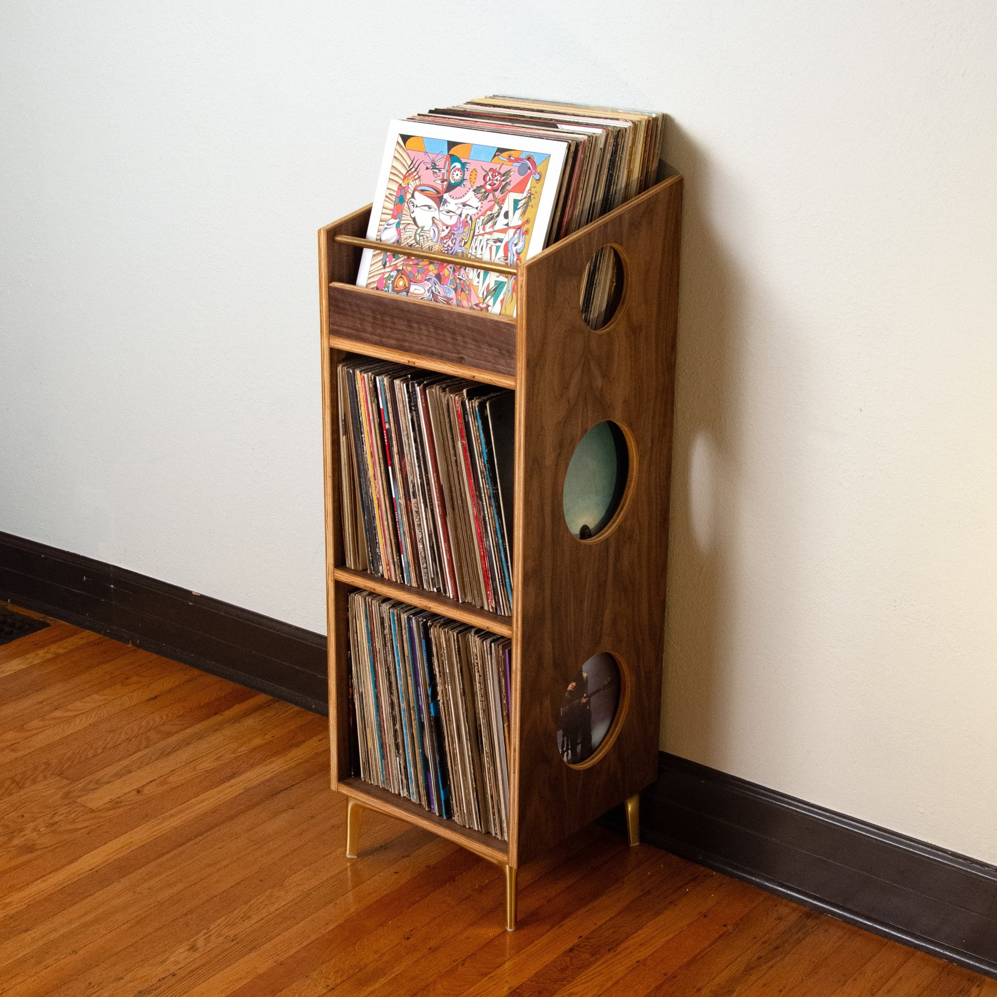 A wooden record rack shelf with circular cutouts, filled with vinyl records, standing on a hardwood floor against a plain off-white wall.