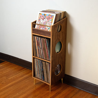 A wooden record rack shelf with circular cutouts, filled with vinyl records, standing on a hardwood floor against a plain off-white wall.