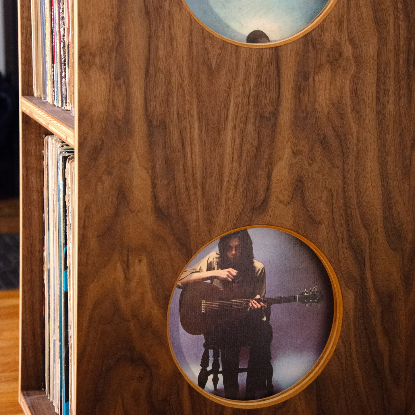 A close-up of a wooden cabinet or record shelf with two circular cutouts showing printed images inside, including a vinyl record sleeve on the left edge.