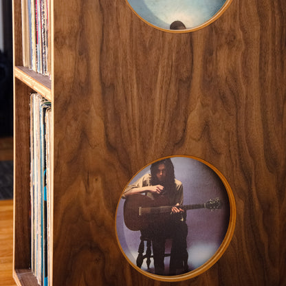A close-up of a wooden cabinet or record shelf with two circular cutouts showing printed images inside, including a vinyl record sleeve on the left edge.