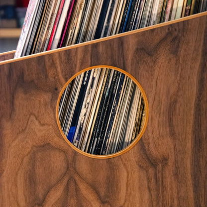 Close-up of a wooden record rack or shelf with circular cutouts showing vinyl records inside, set against a warm wood-grain surface.