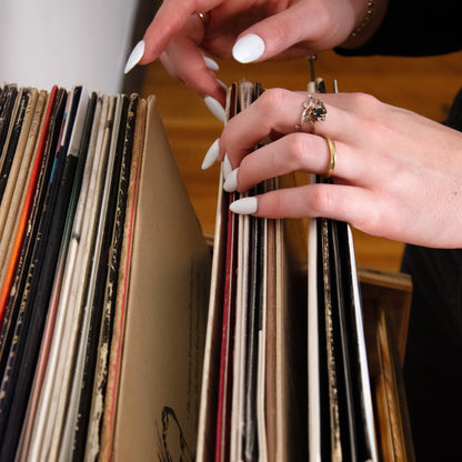 A close-up lifestyle shot of a person's hands with white manicured nails browsing through a stack of vintage vinyl record sleeves on a wooden shelf.