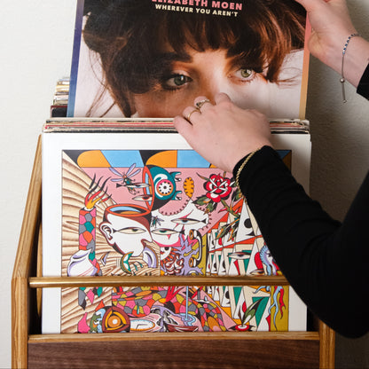 A person's hands holding an album cover for Elizabeth Moen's 'Wherever You Arent' above a wooden record rack filled with other colorful records against a plain off-white wall.