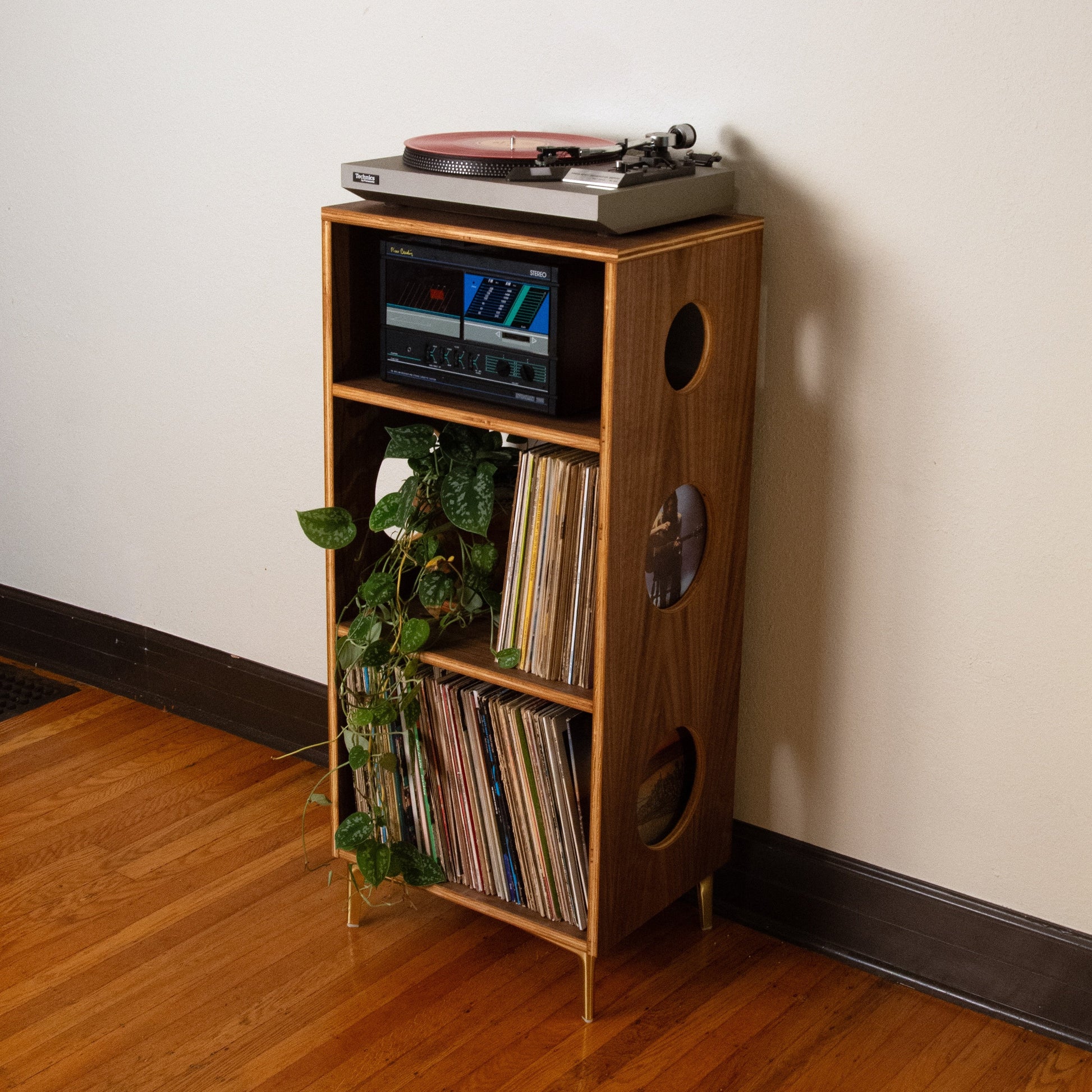 A mid-century modern wooden record player shelf with gold legs, styled with a turntable, records, and a small green plant, set against a plain off-white wall on a hardwood floor.