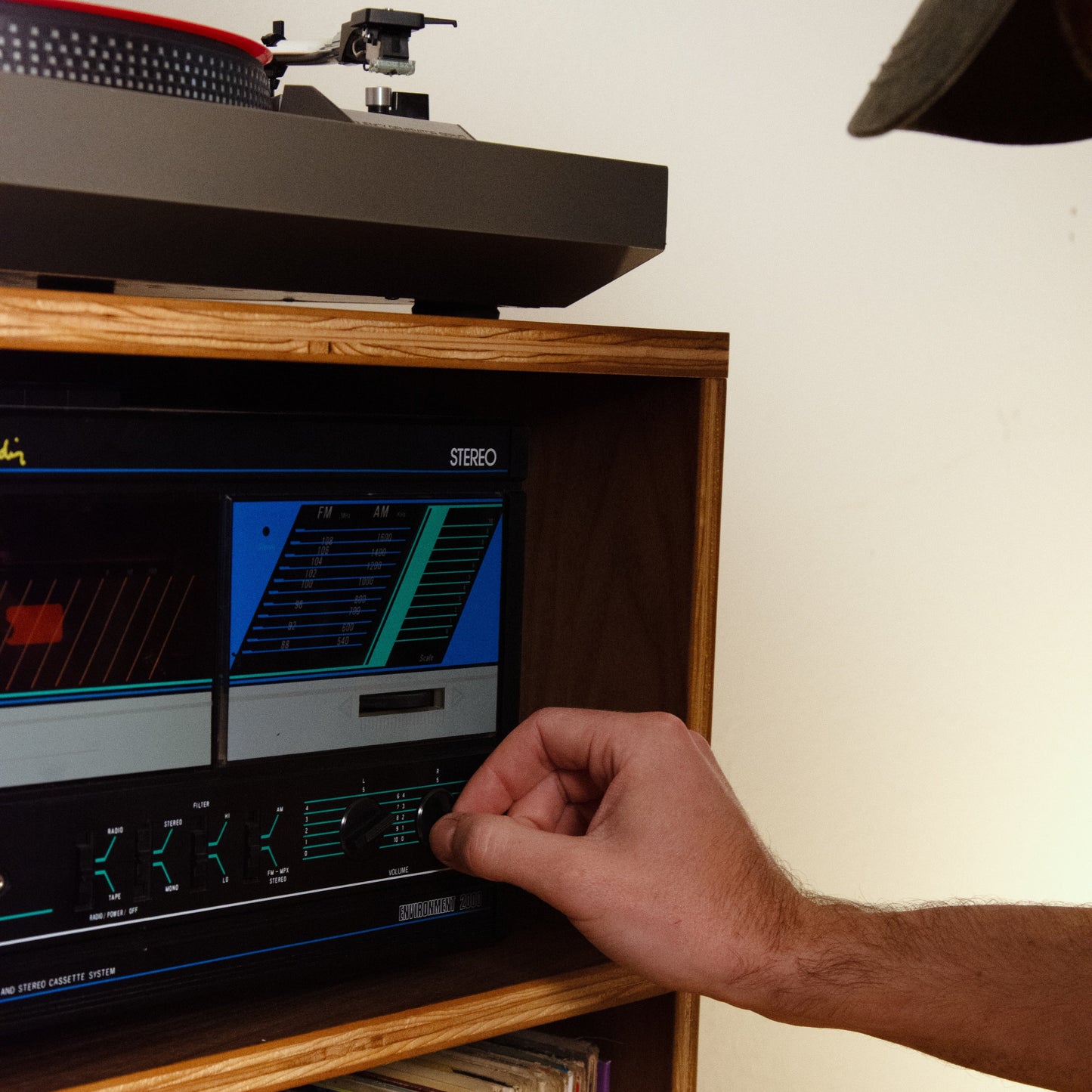 A close-up lifestyle shot of a person's hand adjusting the controls on a vintage Steinway audio receiver inside a wooden shelf cabinet, with a turntable on top and vinyl records below.