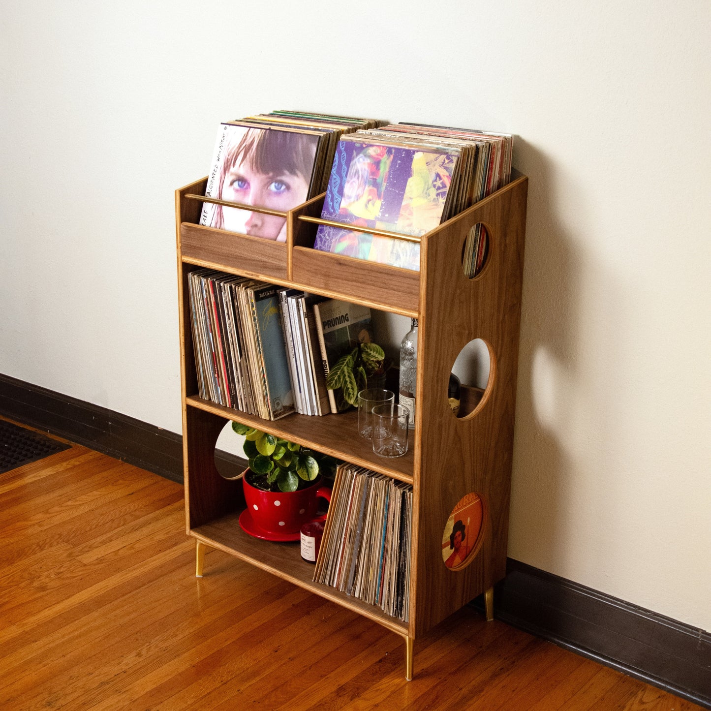 A wooden record shelf with three tiers and side cutouts, filled with vinyl records, standing on a hardwood floor against a plain wall.