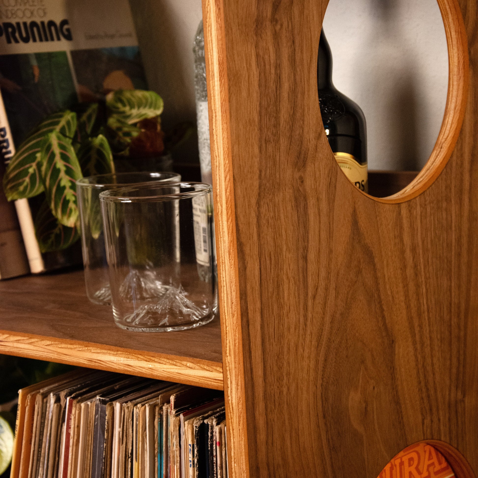 A close-up of a warm medium-brown wooden record shelf with a circular cutout, holding glass tumblers, a magazine, and a plant, with vinyl records visible in the lower shelf.
