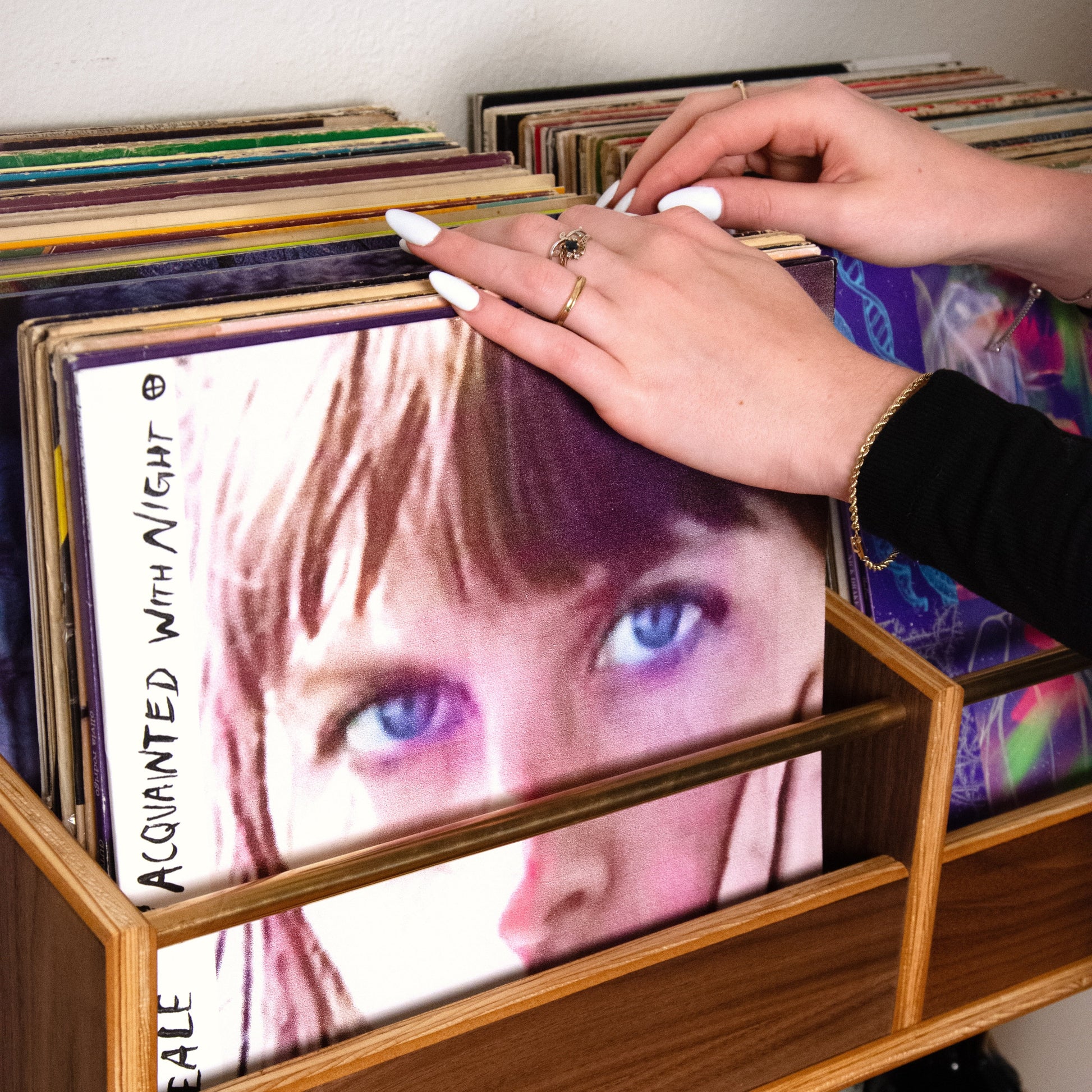 A wooden record shelf filled with vinyl records, with a person's hands placing a record cover featuring a close-up portrait of a woman with blue eyes.